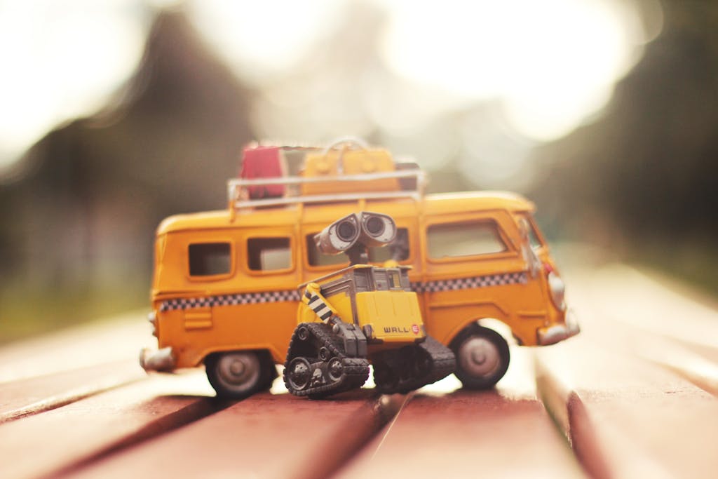 Charming miniature robot with yellow van toy in blurred outdoor daylight, capturing playful leisure theme.