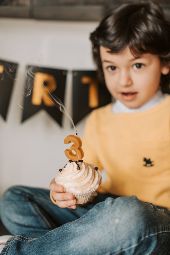 Young child holding a cupcake with number 3 candle during a birthday celebration indoors.